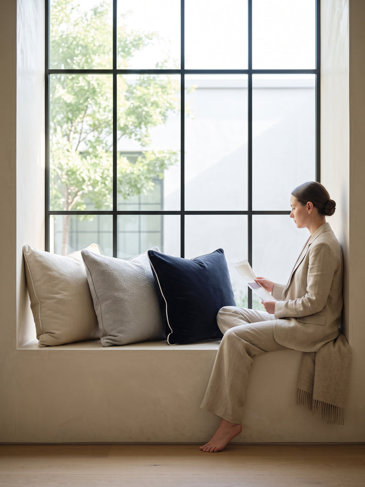 A woman reading on a window seat among bone, grey and navy pillows